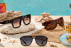 Assorted sunglasses on a sandy beach with driftwood and seashells, turquoise water in the background.