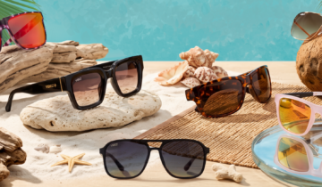 Assorted sunglasses on a sandy beach with driftwood and seashells, turquoise water in the background.